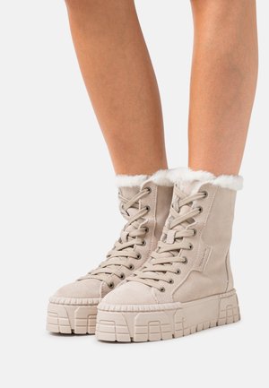 Bottines en suède beige à lacets avec des semelles texturées épaisses et une bordure en fourrure blanche, portées sur des jambes nues sur un fond blanc.