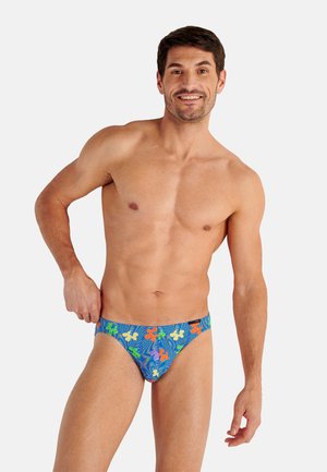 Lachende man met blauwe zwembroek met kleurrijke bloemenpatronen, staand tegen een effen lichte achtergrond.