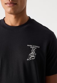 Melna kokvilnas T-kreklu ar baltu grafiku, kurā attēlots lācis, kurš spēlē ģitāru, ar tekstu "MARC O'POLO DENIM" dizaina augšpusē. Klasisks apļa izgriezums.