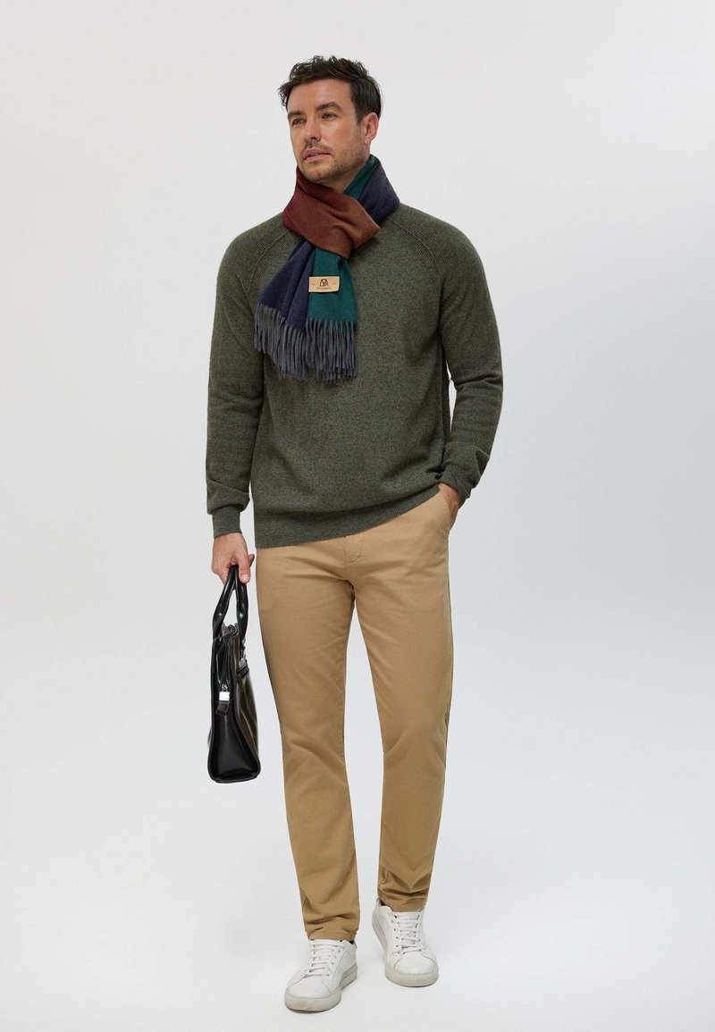 Pull vert olive, pantalon beige et baskets blanches. Une écharpe multicolore drape autour du cou, avec des franges. Sac à main noir à la main.