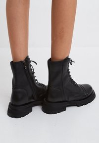 Sorte læderkampstøvler med snørebånd foran, polstret krave og lynlås i siden. Tykk sort gummisål med struktureret slidbane.