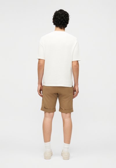 Άνδρας με κατσάρα μαλλιά που φοράει λευκό πουκάμισο με κοντά μανίκια, καφέ ξεστραβωμένες βερμούδες, λευκές κάλτσες και λευκά αθλητικά παπούτσια, στέκεται γυρισμένος με την πλάτη προς εμάς σε μονόχρωμο φόντο.