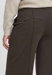 Pantalons marron sur mesure en tissu texturé. Les caractéristiques comprennent des poches latérales, une coupe élégante et des passants de ceinture. Les coutures visibles ajoutent du détail.