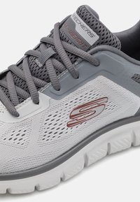 Gray and white athletic shoe with mesh upper, textured detailing, laces, and a prominent logo. Lightweight cushioning and structured sole.