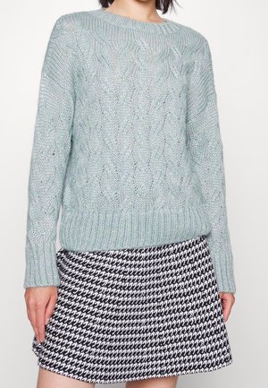Personne portant un pull en maille torsadée bleu clair et une jupe à motif pied-de-poule noir et blanc, debout devant un fond uni.