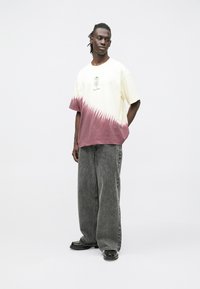 Krótka koszulka z krótkim rękawem w jasnym żółtym kolorze z różowym wzorem dip-dye na dole i kwiatową grafiką z przodu, zestawiona z szerokimi szarymi dżinsami.