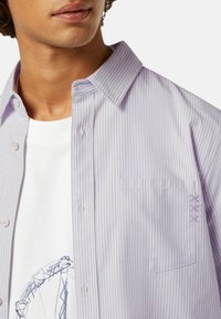 Chemise rayée lavande claire avec col à boutons, dotée d'une poche poitrine et de coutures décoratives violettes sur la manche.