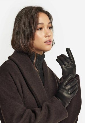 Gants en cuir noir à finition lisse, présentant un design ajusté et trois coutures bien distinctes pour les doigts, portés par une personne dans un manteau marron.