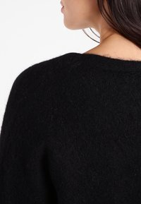 Černý, chlupatý svetr s širokým výstřihem a volným střihem. Jemná textura s mírným leskem, která představuje minimalistický design.