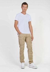 Jonge man staat met zijn handen in zijn zakken, draagt een witte V-hals T-shirt, beige cargobroek en witte sneakers tegen een effen achtergrond.