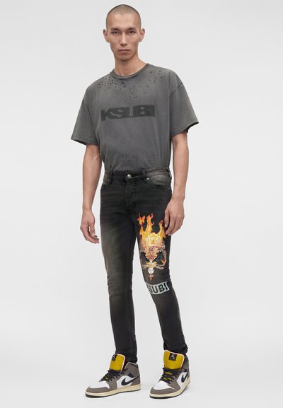 Άνδρας με ξυρισμένο κεφάλι που φοράει σκούρο γκρι φθαρμένο κοντομάνικο Ksubi, μαύρο τζιν με γραφικό σχέδιο κρανίου με φλόγες και γκρι ψηλά αθλητικά παπούτσια Nike.