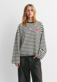 Jonge vrouw draagt een zwart-wit gestreept shirt met lange mouwen en rode tekst, gecombineerd met donkere, loszittende jeans, staand tegen een effen witte achtergrond.