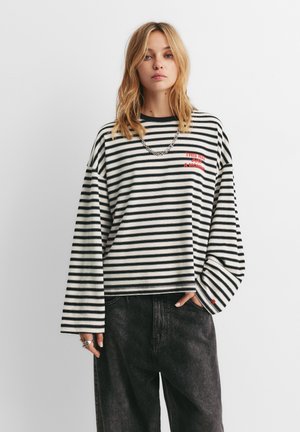 Jonge vrouw draagt een zwart-wit gestreept shirt met lange mouwen en rode tekst, gecombineerd met donkere, loszittende jeans, staand tegen een effen witte achtergrond.