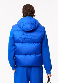 Modrá puffa vesta s kapucňou, s lesklým povrchom, prešívaným dizajnom a vyšitým logom na chrbte, doplnená o zodpovedajúci modrý outfit.