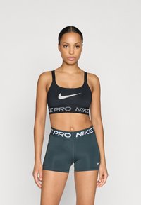 Czarny stanik sportowy z białym logo Nike i teksturowanym pasem. Ciemnozielone spodenki z pasem Nike Pro. Gładki, elastyczny materiał, dopasowany krój.