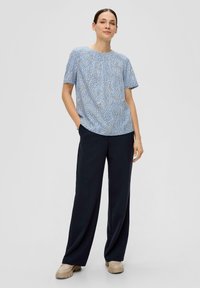 Haut à manches courtes bleu à motifs, avec un col rond, associé à un pantalon ample foncé et des chaussures beiges. Texture lisse, design décontracté.