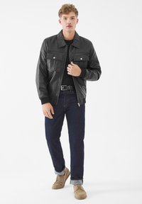 Junger Mann in schwarzer Lederjacke, schwarzem Hemd, dunkelblauer Jeans mit Umschlägen und hellbraunen Schuhen, der vor einem schlichten weißen Hintergrund steht.