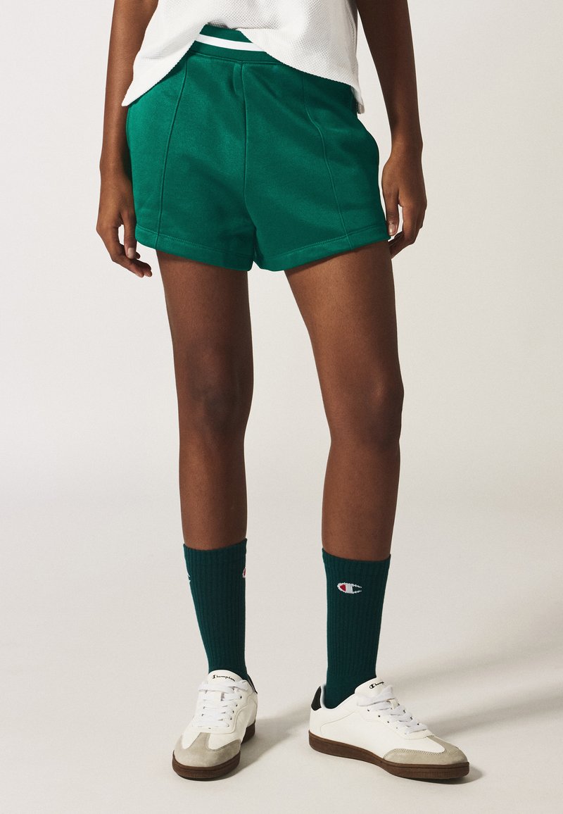 Pantalones cortos verdes con una textura suave y cinturilla elástica, combinados con calcetines verde oscuro y zapatillas blancas con detalles grises.