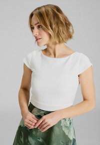 Top bianco aderente con maniche corte, scollatura rotonda e texture liscia, abbinato a una gonna verde con motivo floreale. Focus su linee pulite.