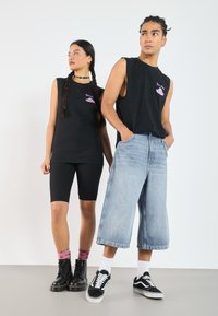 Schwarze ärmellose T-Shirts mit grafischem Design, kombiniert mit schwarzen gerippten Shorts und weiten Jeans-Culottes. Beide tragen kontrastierende Schuhe.