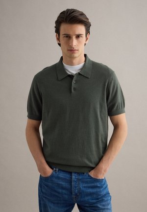 Jonge man met donker haar, gekleed in een donkergroen poloshirt met korte mouwen over een wit shirt, handen in de zakken, staand tegen een effen achtergrond.