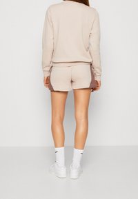 Beżowa bluza z długimi rękawami, zestawiona z pasującymi szortami z brązowymi bocznymi akcentami. Dopełnieniem stroju są białe sneakersy i skarpetki.