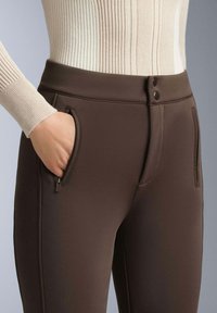 Hnědé elastické kalhoty s hladkou texturou, vybavené bočními kapsami na zip a knoflíkovým zapínáním v pase. Vrchní část má žebrovaný design.