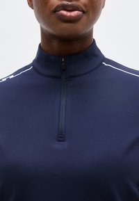 Κοντινό πλάνο του κάτω μέρους του προσώπου και του λαιμού ενός ατόμου που φοράει μπλε ναυτικό αθλητικό τοπ με φερμουάρ στο λαιμό και λευκές ρίγες στους ώμους.