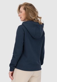 Felpa con cappuccio blu navy dal tessuto morbido, con polsini a costine e cappuccio regolabile, indossata sopra pantaloni chiari.