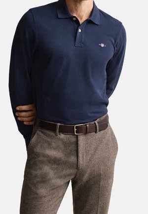 Man wearing navy long-sleeve polo shirt tucked into brown herringbone trousers with dark brown belt, standing with one arm behind back.