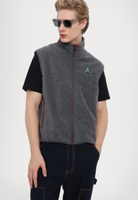 Gray fleece vest with a high collar, red zipper accents, and a blue embroidered logo, worn over a black t-shirt and navy pants.