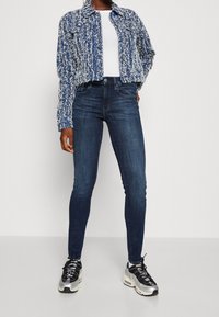 kort blå och vit texturerad jacka över en vit topp, kombinerad med mörkblå skinny jeans och svarta silver sneakers.