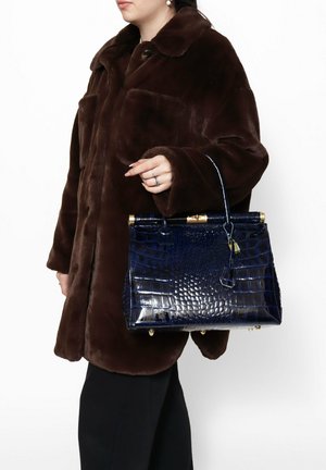 Crocodile-embossed navy handbag with a structured shape, gold hardware accents, and a top handle, worn with a brown faux fur coat.