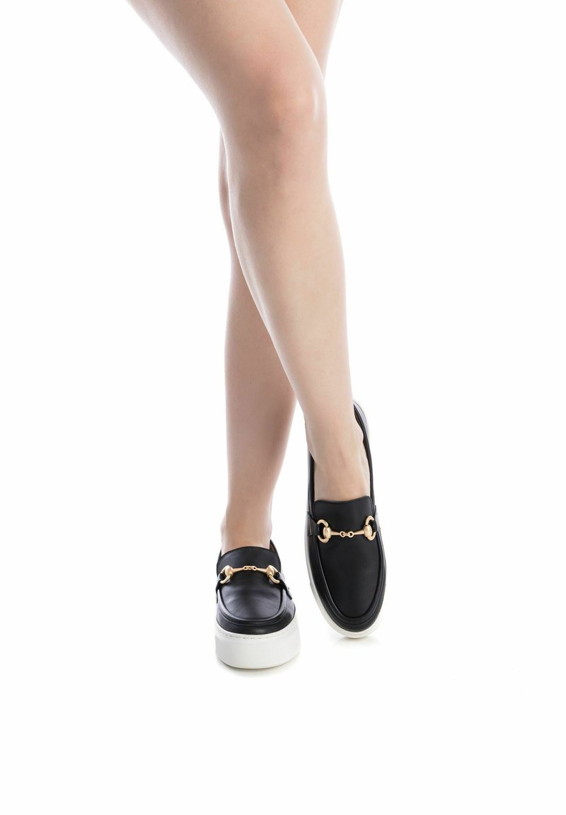 Zapatos negros de cuero tipo slip-on con acento de cadena dorada, suela plataforma blanca, puntera redondeada, textura suave y detalles de diseño mínimos.