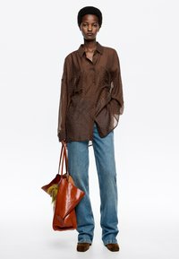 Camisa marrón semitranslúcida con botones delanteros y lunares, combinada con jeans azul claro, sosteniendo un bolso de cuero naranja y zapatos marrones.