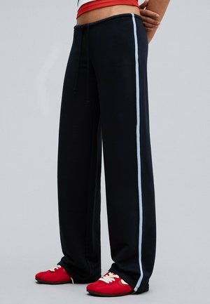 Personne portant un pantalon noir à jambes larges avec des bandes latérales bleu clair et des baskets rouges, debout devant un fond gris uni.