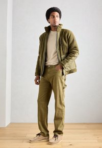 Olivgrüne Fleecejacke mit hohem Kragen, khakifarbenes T-Shirt und passende Cargo-Hosen. Beigefarbene Schuhe mit braunen Schnürsenkeln, stehen auf einem Holzboden.