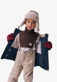 Enfant portant un pull en laine tricoté marron, un pantalon de neige beige et des gants rouges. Il porte un bonnet doublé en peluche et tient une veste ouverte bleu marine et beige.