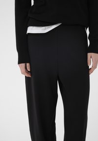 Črne široke hlače iz mehke tkanine, z ravnim sprednjim delom, subtilnim stranskim šivom, kombinirane z belo večplastno srajco pod njimi.