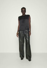 Mulher a usar uma blusa preta sem mangas, calças largas de pele preta, sapatos pretos de bico fino, óculos de sol grandes e quadrados, e brincos e pulseira de argola dourada.