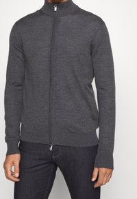 Man som bär en mörkgrå zip-up tröja med ribbade ärmslut och fåll, tillsammans med mörkblåjeans.