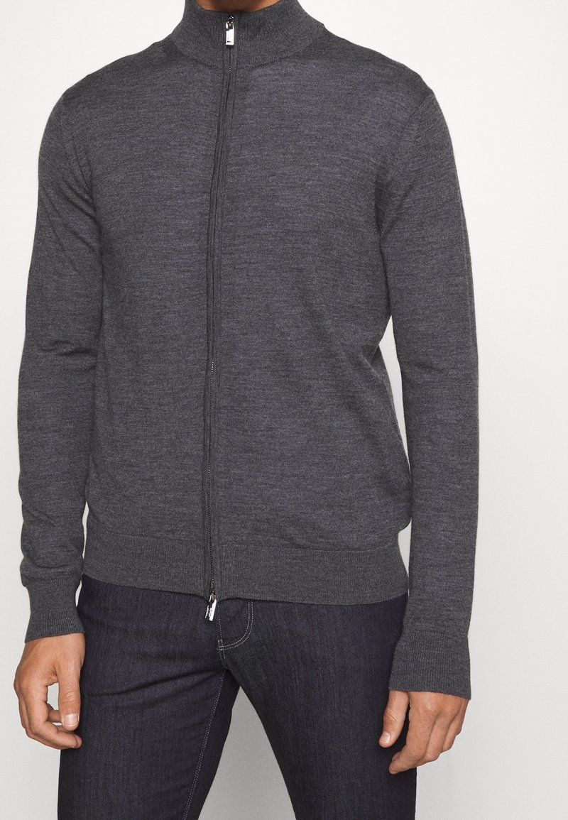 Man som bär en mörkgrå zip-up tröja med ribbade ärmslut och fåll, tillsammans med mörkblåjeans.