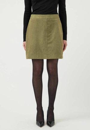 Woman wearing a knee-length olive green skirt, black sheer tights, black pointed-toe heels, and a long-sleeve black top against a white background.