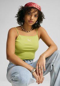 Canottiera verde a costine con spalline sottili e logo bianco, abbinata a jeans leggeri azzurri a vita alta. Gli accessori includono un bandana rossa e gioielli.