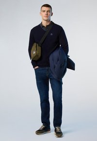 Dunkelblauer V-Ausschnitt-Pullover über einem Hemd mit Kragen, kombiniert mit dunklen Jeans und Sportschuhen. Eine blaue Steppjacke und eine olivgrüne Umhängetasche werden gehalten.