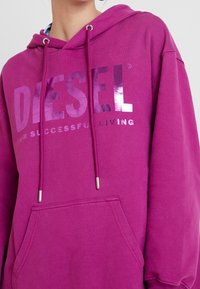 Kvinna som bär en magenta huvtröja med en glänsande "DIESEL"-logotyp och dragsnores, med en ficka framtill.