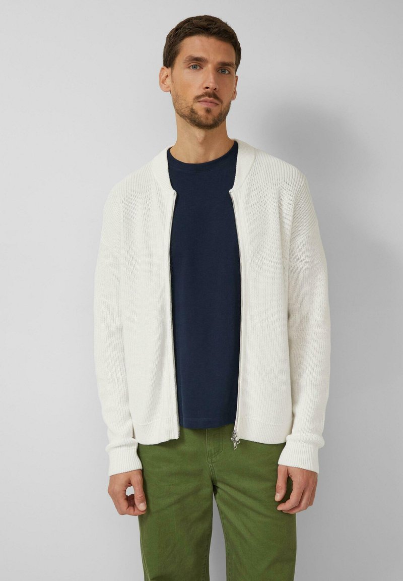 Hellbeiger Reißverschluss-Cardigan mit gerippter Struktur, rundem Kragen und langen Ärmeln, über einem marineblauen Hemd und grüner Hose getragen.