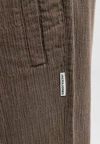 Tissu marron texturé avec une étiquette blanche cousue portant la mention « JACK & JONES » près d'une couture verticale et d'un petit bord de poche.