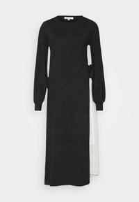 La robe noire à manches longues présente une texture côtelée, une silhouette décontractée et un accent superposé blanc noué à la taille, créant un contraste de couleurs.