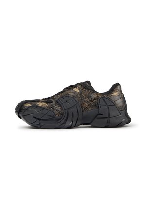 Schwarz-goldener, abgenutzter Sneaker mit dicker segmentierter Sohle und strukturiertem Obermaterial, seitlich auf weißem Hintergrund abgebildet.
